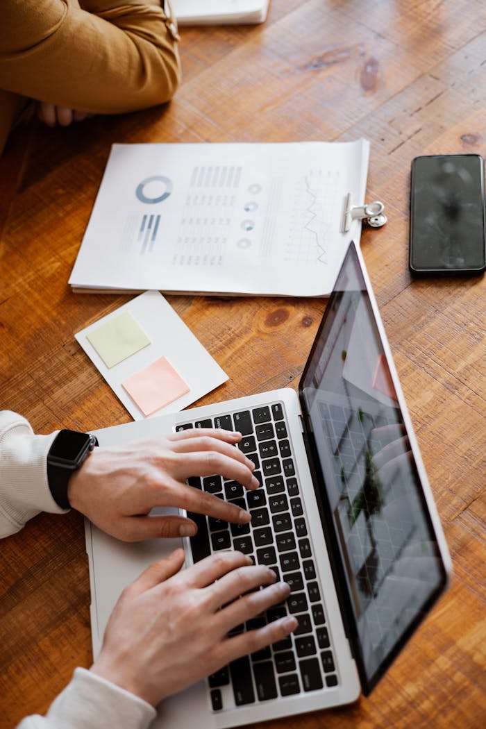 Hands typing on a laptop with paperwork and smartphone on a wooden desk, ideal for business and office themes.